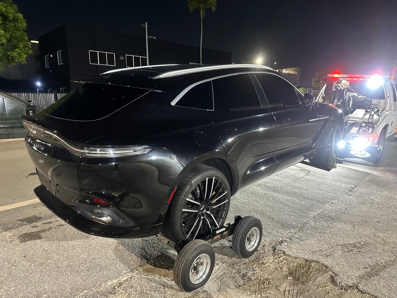 Black Aston Martin DBX SUV at night, rear wheels raised on tow dollies with the lit wrecker parked behind it on a Miami street