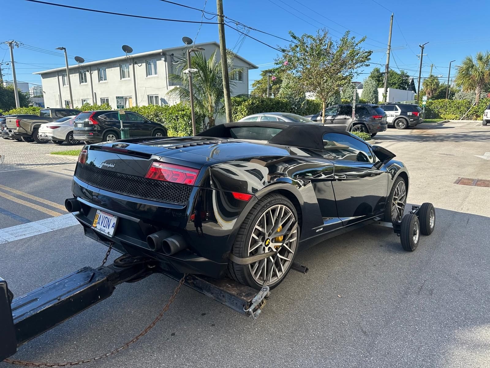 Black Lamborghini Gallardo Spyder on tow dollies on a Miami street in daylight, wrecker attached